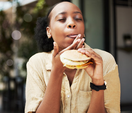 Hamburger, fast food and black woman eating a brunch in an outdoor restaurant as a lunch meal craving deal. Breakfast, sandwich and young female person or customer enjoying a tasty unhealthy snackの写真素材
