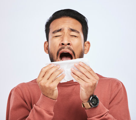 Allergy, sneeze and asian man is sick with tissue in studio with grey background with cold or sinus. Virus, allergies and male person with toilet paper for flu or problem with nose or congestion.の写真素材