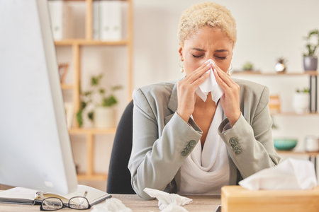 Computer, tissue and a business woman blowing nose while working at a desk, sick in the office. Cold, flu or symptoms with a young female corporate employee sneezing from hayfever allergies at workの写真素材