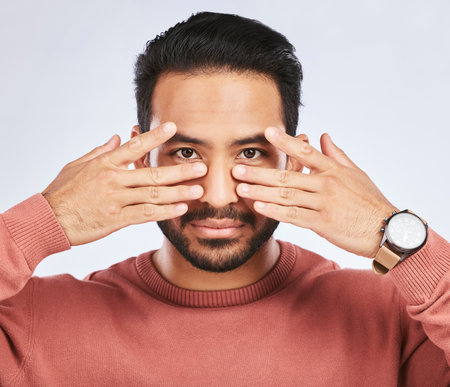 Portrait, eyes and asian man with fingers on face in studio with confidence, personality and persona on grey background. Hands, pose and male person with hand gesture for mask, masquerade or egoの写真素材