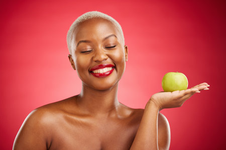 Happy black woman, apple and natural nutrition for healthy diet against a red studio background. African female person smile with natural organic green fruit for food snack, health and wellnessの写真素材