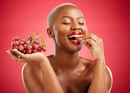 Health, eating and a black woman with grapes on a red background for nutrition or diet. Smile, beauty and an African model or girl with fruit, hungry and food for a detox isolated on a backdropの写真素材