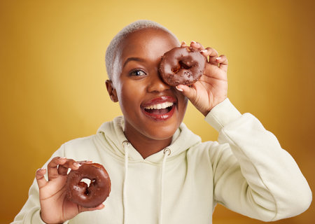 Portrait, chocolate and donut with a black woman in studio on a gold background for candy or unhealthy eating. Smile, food and baking with a happy young female person holding sweet pastry for dessertの写真素材
