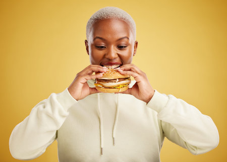 Burger, eating and woman or happy student on a studio, yellow background for restaurant promotion or...