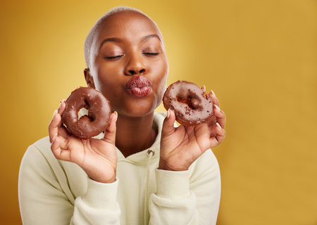 Face, kiss and donut with a black woman in studio on a golden background for candy or unhealthy eating. Pout, food and baking with a cute young female person holding sweet pastry for dessertの写真素材