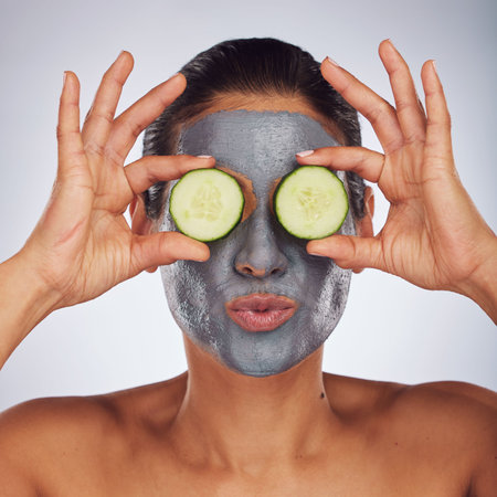 Eyes, cucumber and mask with a model woman in studio on a gray background for an antiaging facial treatment. Face, skincare and food with a young female person using cosmetic clay on her faceの写真素材