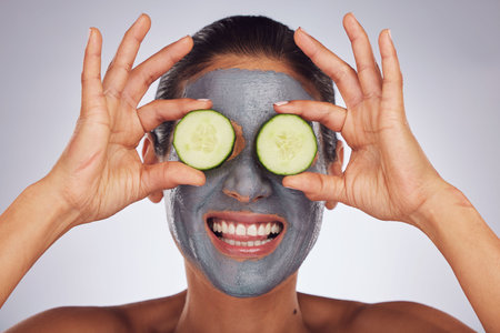 Facial, cucumber and mask with a model woman in studio on a gray background for an antiaging face treatment. Eyes, skincare and food with a happy young female person using cosmetic clay on her faceの写真素材