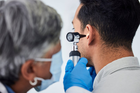 Woman, doctor and checking patient ear for flu, exam or healthcare appointment at the hospital. Female person or medical professional with protection looking at ill or sick man with flu at the clinicの写真素材