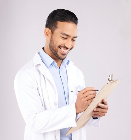 Writing, smile and happy man doctor with checklist in studio for medical, compliance or insurance on grey background. Healthcare, form and male health expert check paperwork or agenda schedule notesの写真素材