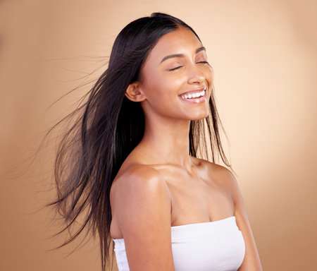 Salon, hairstyle and indian woman with beauty or wind in brown studio background. Happy, girl and haircare with eyes closed or keratin treatment for shine or volume and natural cosmetics with glow.の写真素材