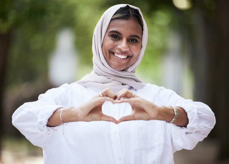 Hands in heart, park and portrait of Muslim woman for support, love symbol and care outdoors. Happy, emoji and face of Islamic female person with hijab and hand sign for kindness or peace in natureの写真素材