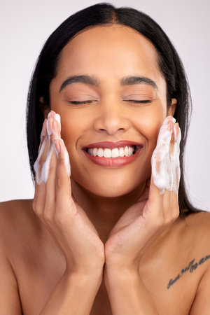 Happy woman, face and washing for skincare hygiene, cleaning or dermatology against a grey studio background. Female person with smile and soap hands for clean facial treatment, self care or loveの写真素材