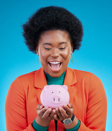 Woman, piggy bank and wow for savings, financial investment and profit, increase or budget success. Excited african person with banking goals, finance safe and container on a blue, studio backgroundの写真素材