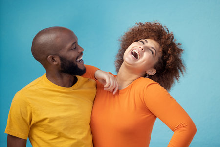 Love, laughing and interracial couple with a joke isolated on a blue background in a studio. Comic, funny and black man and woman smiling with happiness, care and confidence on a backdrop togetherの写真素材