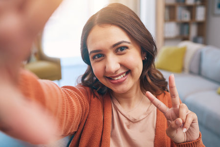 Portrait, woman and peace for selfie in home, smile and fun with live streaming vlog in living room. Face, profile picture and happy female gen z show v sign, hand emoji and memory for social mediaの写真素材