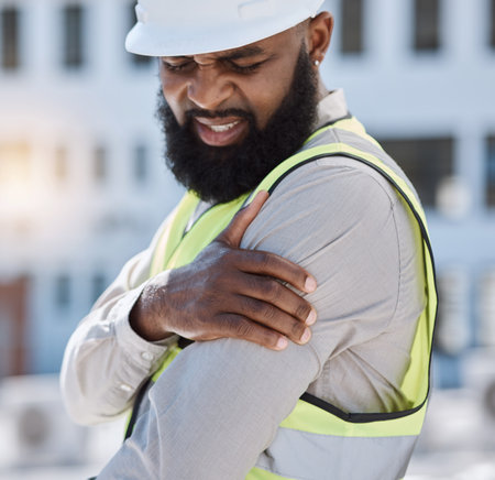 Injury, arm pain and a black man construction worker on a building site, feeling fatigue in his shoulder. Burnout, emergency or accident and a male engineer holding his muscle with inflammationの写真素材