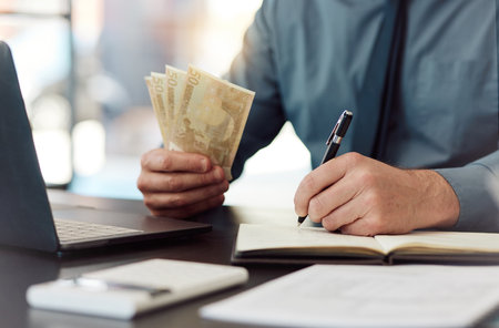 Hands, money and saving with writing, book and accounting for audit, balance and financial review in workplace. Man, pen and notebook with cash, bills and budget for small business, investing or goalの写真素材