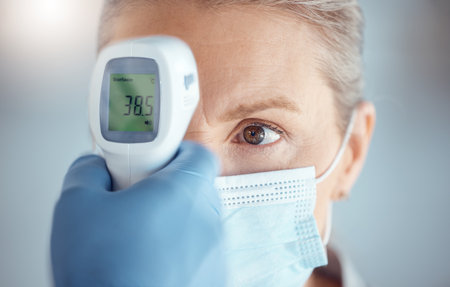 Woman in mask, hand of doctor and digital thermometer in hospital for covid, safety and healthcare consultation. Temperature test, corona and patient at clinic with medical professional for exam.の写真素材