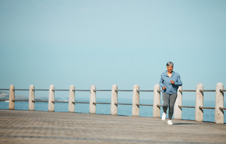 Fitness, running and senior woman at the beach for exercise, health or cardio on blue sky background. Training, wellness and elderly female runner at the sea for morning workout, routine or ocean runの写真素材