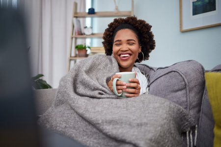 Laptop, coffee and woman with blanket on sofa laughing for comedy, funny and comic movies. Happy, home and African female person relax with computer for entertainment, streaming and watching filmの写真素材