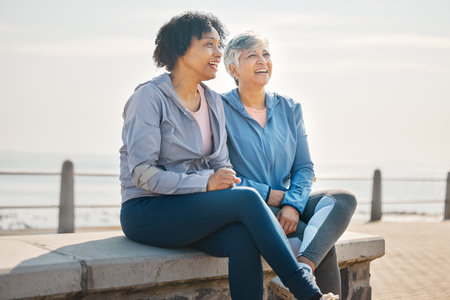 Senior women, sitting and beach with friends, training and laugh in summer outdoor. Exercise, break and mature female in the morning with funny joke and workout for wellness and health by seaの写真素材