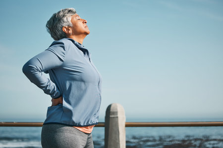 Tired, running and senior woman pain from back ache by the ocean for fitness, exercise and workout....