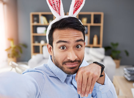 Bunny ears, holiday and selfie with a man and remote work on easter with creative job. Celebration, happy and male professional from Spain feeling silly and goofy with comedy rabbit hat in a homeの写真素材