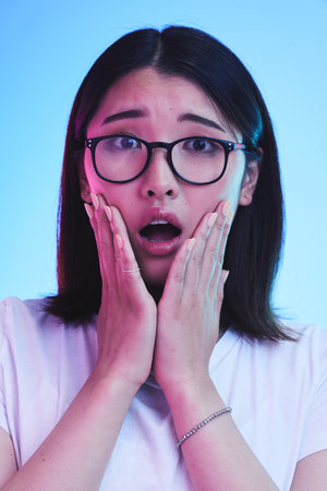 Surprise, shocked and face of Asian woman with wow expression or open mouth for drama, deal and promotion. Omg, wtf and portrait of person with discount emoji isolated in a studio blue backgroundの写真素材