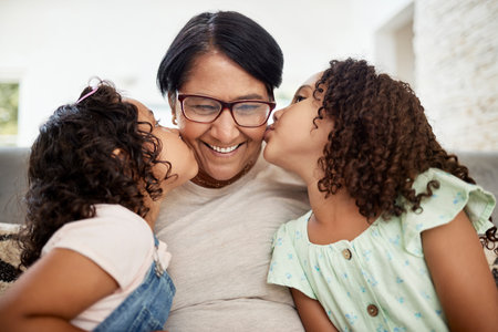 Kids kissing their grandmother on the cheek with care, love and happiness while relaxing in the living room. Smile, happy and senior woman hugging girl children for bonding together at family home.の写真素材