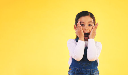 Portrait, fashion and hands on face of girl child in studio with deal, sale or giveaway promo on yellow background. Excited, surprise and open mouth kid with emoji for news, announcement or wow infoの写真素材