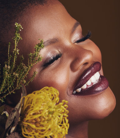Face, beauty and protea with the smile of a black woman in studio on brown background for natural treatment. Happy, plant or cosmetics and a young model indoor for aesthetic wellness with flowersの写真素材