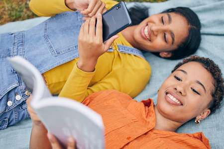 Girl friends, park and top view with reading, book and smartphone for blog, relax or knowledge in nature. Gen z students, phone and story on blanket, picnic or outdoor for holiday, backyard or gardenの写真素材