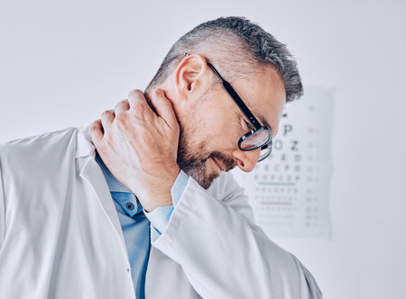Healthcare, neck pain or optometry and a doctor in his office with an injury while feeling stress. Medical, burnout or accident with a mature optician holding his spine or shoulder in a clinicの写真素材