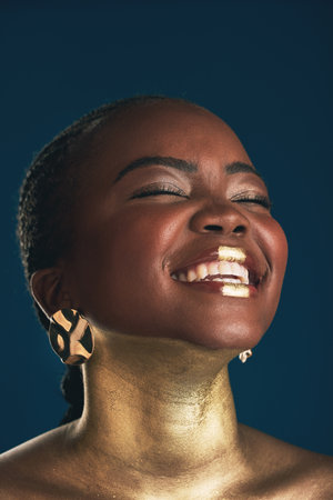 Gold makeup, face and black woman laughing at funny creativity, facial cosmetics paint and beauty art design. African culture joke, comedy or glamour person with creative face glow on blue backgroundの写真素材