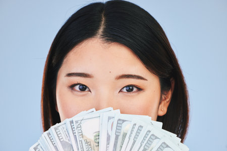 Money, beauty and woman cover face on blue background for winning, cash fan or financial prize. Lottery, portrait and asian person or winner with bonus, cosmetics secret or finance profit in studioの写真素材
