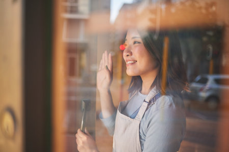 Asian woman, window and door of restaurant to welcome service, small business owner and wave with reflection. Hello from manager, smile and Japanese food cafe startup with happy entrepreneur at work.の写真素材