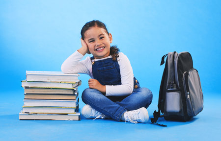 Girl child, sitting and books in studio portrait, backpack and excited for learning on floor by blue background. Female kid, education and development with smile, future and studying for knowledgeの写真素材