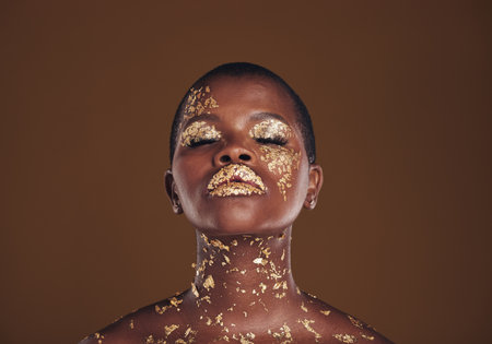 Beauty, glitter and black woman with gold makeup on brown background with art, paint and cosmetics. Shine, glow and African model in studio for facial fashion, aesthetic freedom and luxury skincare.の写真素材