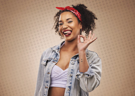 Woman, ok sign and studio portrait with smile, tongue and funny face for good review by brown background. African girl, student or fashion model with hand for icon, vote or feedback with comic laughの写真素材
