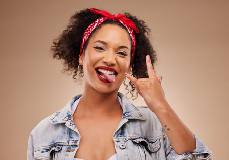 Woman, horns sign and studio portrait with tongue, comic smile and funny face with beauty by brown background. African girl, model or student with icon, crazy and rock star hand with laugh for beautyの写真素材