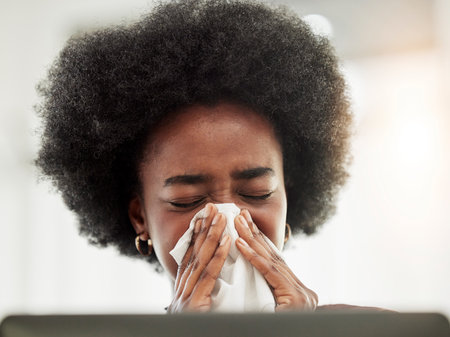 Allergies, sneeze and sick black woman blowing nose in home office with allergies, flu or cold in her home. Tissue, paper and African female freelancer with virus, hayfever or covid working on laptopの写真素材