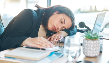 Tired, work and a woman at a desk for sleeping, corporate or working burnout in an office. Narcolepsy, table and a female business employee with a nap, rest or fatigue from company stress or jobの写真素材