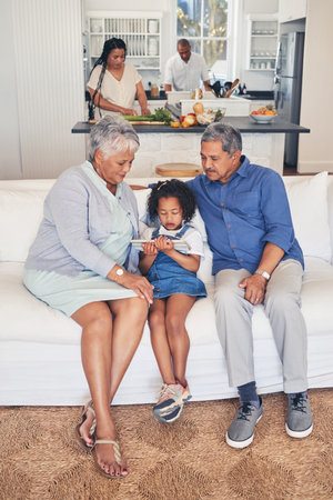 Home, family and grandparents with girl, tablet and weekend break with fun, social media and streaming movies. Granny, old man or kid on a sofa, technology or cartoons with application, film or relaxの写真素材
