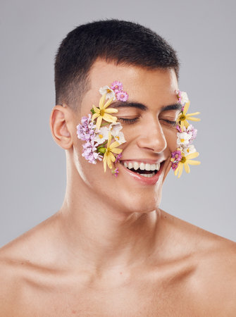 Smile, skincare and a man with flowers on a studio background for creative aesthetic. Happy, plant and a male model with floral art for facial wellness and dermatology isolated on a studio backdropの写真素材