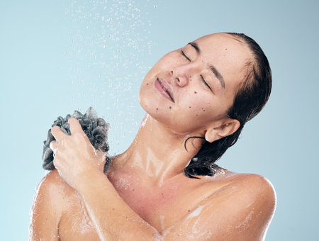 Woman, shower and water drops with loofah in hygiene, grooming or washing against a blue studio background. Female person in relax for body wash, cleaning or skincare routine under rain in bathroomの写真素材
