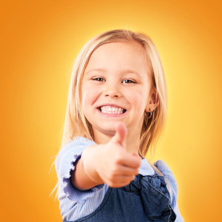 Child, happy portrait and thumbs up in studio for support, like emoji or yes for approval. Face of young girl kid on a orange background for hand gesture, icon or sign for agreement or thank youの写真素材