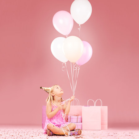 Balloons, birthday and girl on pink background for party, celebration and special day in studio. Happy, celebrate and young child sitting with presents, gift box and decoration for festive eventの写真素材