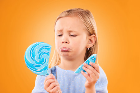 Children, candy and a sad girl with a broken lollipop on an orange background in studio looking...