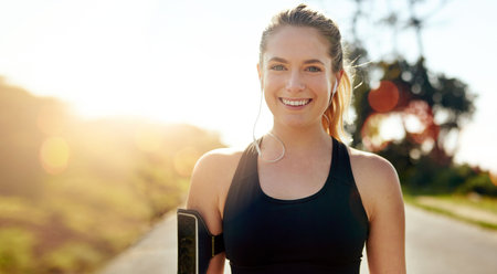 Fitness, portrait and happy woman with earphones outdoor on road at sunrise, training workout or exercise. Listening to music, smile and face of athlete in nature for wellness, healthy body or sportの写真素材