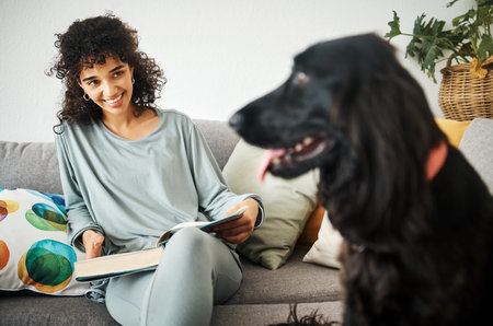 Reading a book, dog and a woman on home sofa to relax with animal in a living room. Pet owner,...